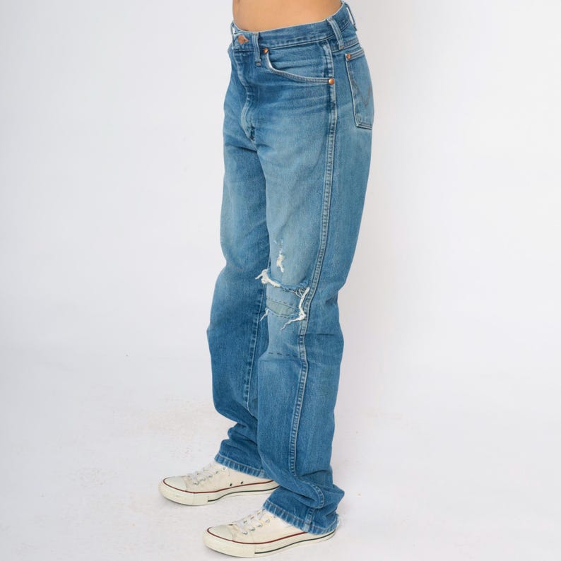 May include: Faded blue denim jeans with distressed patches on the knees. The jeans are worn with white canvas trainers. The overall style is casual and relaxed.