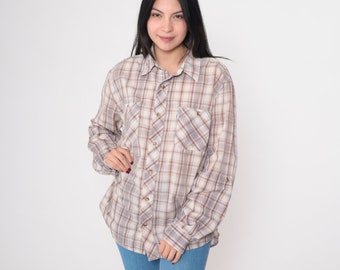 May include: A beige, brown, and red plaid button-down shirt with two chest pockets. The sleeves are rolled up. The shirt is worn open, paired with blue jeans. The shirt is a classic style.