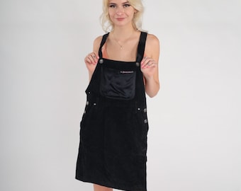 May include: A black corduroy overall dress with a small pocket on the front. The dress has adjustable straps and silver button accents.