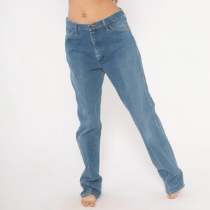 May include: Light blue denim jeans with a classic five-pocket design. The jeans have a straight leg cut and a button and zipper closure. The denim has a faded appearance, with subtle wear marks. The jeans are worn at the waist.