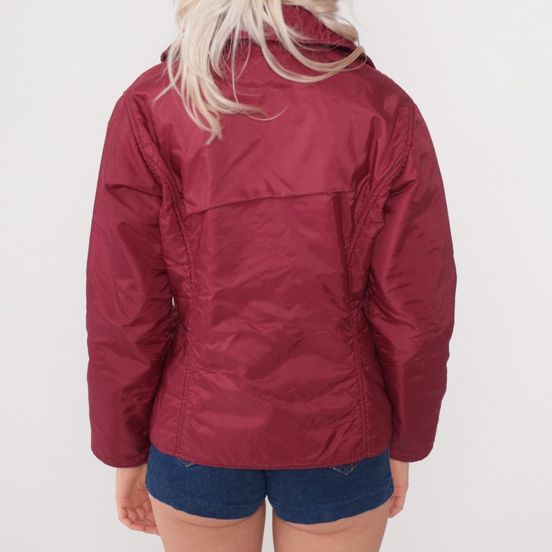May include: A burgundy-colored jacket with a collared neckline and a zipper closure. The jacket has a quilted appearance and a slightly shiny finish. The back view shows a fitted silhouette, with seams running down the sides and back.