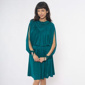 May include: Teal-colored dress with a cowl neckline and open sleeves. The dress has a cinched waist and tie details at the wrists. The model is wearing the dress in front of a white background.