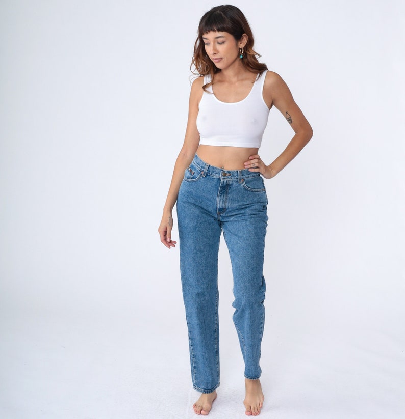 May include: A woman in a white crop top and blue jeans. The jeans are light wash denim with a straight leg. The top is a simple tank style. The background is plain white.
