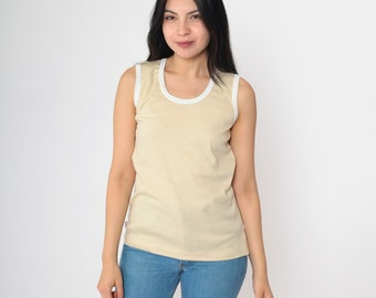 Può includere: Una canotta senza maniche di colore beige chiaro a tinta unita con un bordo bianco a contrasto attorno allo scollo rotondo e agli scalfi, abbinata a jeans blu. Il capo presenta un design semplice a taglio dritto con finitura pulita.