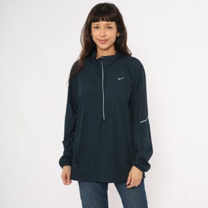Könnte beinhalten: Dunkelblau-grüne Nike-Pulloverjacke mit Kapuze und langen Ärmeln. Die Jacke hat einen weißen Reißverschluss, ein kleines Nike-Logo und weiße Akzente an den Ärmeln. Das Model trägt eine blaue Jeans.