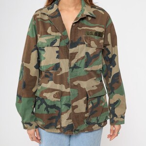 May include: A green, brown, and black camouflage jacket with a button closure and two large pockets on the front. The jacket has a patch on the left chest that says "U.S. Army".