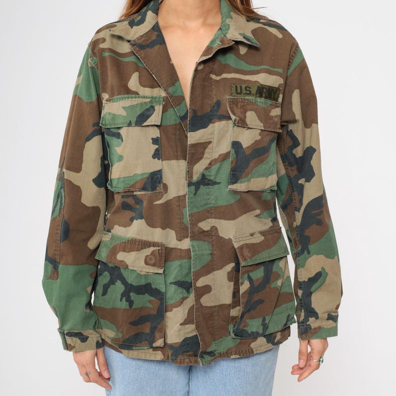 May include: A green, brown, and black camouflage jacket with a button closure and two large pockets on the front. The jacket has a patch on the left chest that says "U.S. Army".
