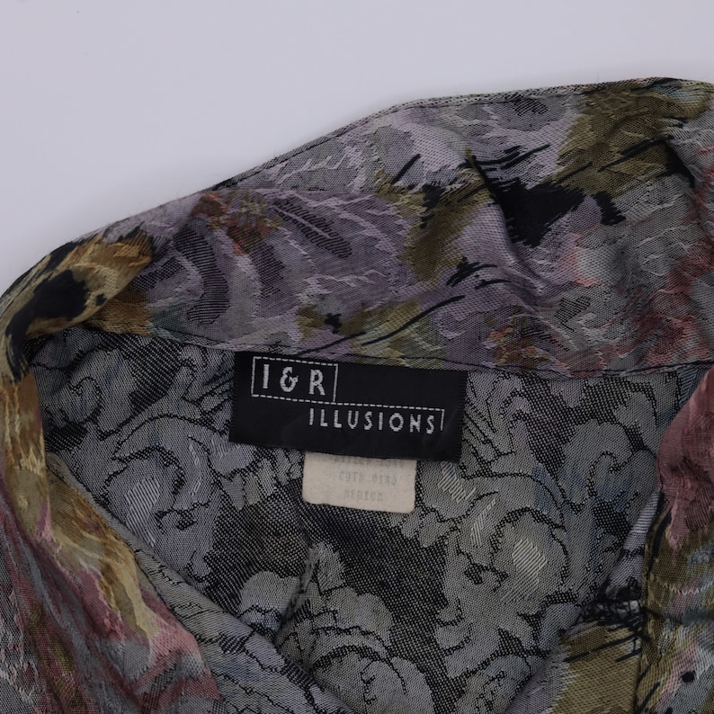 May include: Close-up of a patterned button-down shirt. The fabric features a floral design in shades of grey, green, and pink. The label reads "I&R ILLUSIONS".