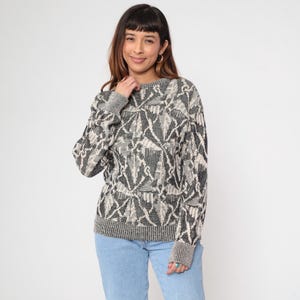 May include: A gray, black, and cream-colored sweater with a geometric pattern. The sweater has a crew neck, long sleeves, and ribbed cuffs and hem. The model is wearing light blue jeans.