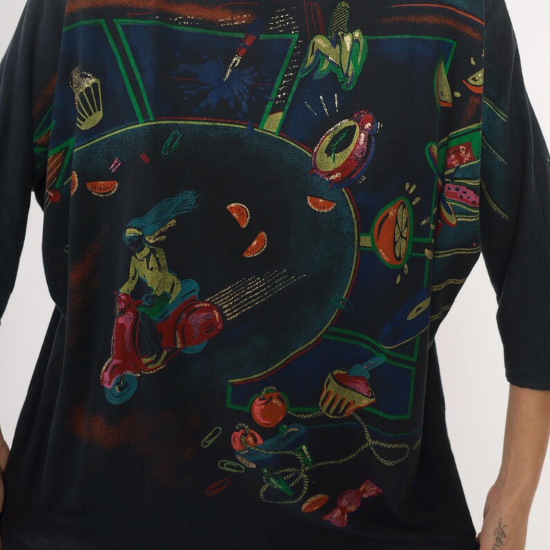 May include: Black long-sleeve shirt with a colorful graphic print. The design features a person riding a red scooter, surrounded by fruit and other decorative elements. The artwork has a retro, vintage style with bright colors.