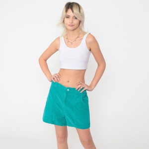 May include: A white sleeveless crop top paired with high-waisted teal corduroy shorts. The shorts feature a front button closure, two side pockets, and vertical seam detailing down the front panels, creating a structured and textured appearance.