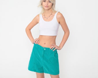 May include: A white sleeveless crop top paired with high-waisted teal corduroy shorts. The shorts feature a front button closure, two side pockets, and vertical seam detailing down the front panels, creating a structured and textured appearance.