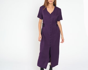 May include: A deep purple, button-down dress with short sleeves and a V-neck. The dress has a fitted waist and a slit up the front. The model is wearing black boots. The dress is made of a smooth fabric.