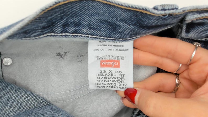 May include: Close-up of a pair of blue denim jeans. The interior label reads "Wrangler" with details including size 33 x 30, relaxed fit, and fabric composition. The jeans are made in Mexico and are 100% cotton.