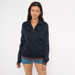 May include: A navy blue zip-up track jacket featuring a high collar, long sleeves, and ribbed cuffs. The garment includes horizontal panel detailing across the chest and a hemline accented with thin red and white stripes. The fabric has a smooth finish.