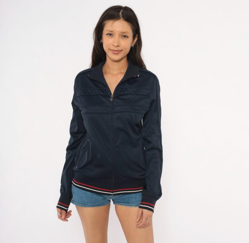 May include: A navy blue zip-up track jacket featuring a high collar, long sleeves, and ribbed cuffs. The garment includes horizontal panel detailing across the chest and a hemline accented with thin red and white stripes. The fabric has a smooth finish.