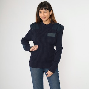 May include: A navy blue ribbed knit jumper with shoulder and elbow patches. The jumper has a crew neck and long sleeves. The patches are a darker shade of navy blue. The person is wearing blue jeans.