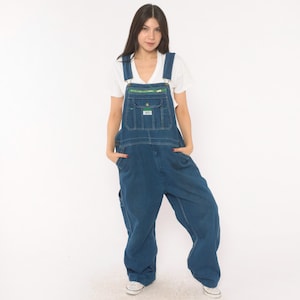 May include: A white short-sleeved t-shirt worn under dark blue denim overalls. The overalls feature a large front bib pocket with a bright green zipper closure, adjustable shoulder straps with metal buckles, and a loose, relaxed-fit leg design.
