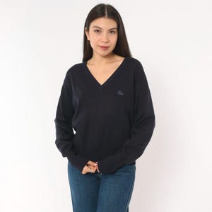 May include: A dark navy blue long-sleeve sweater featuring a V-neckline and a small tonal embroidered logo on the left chest. The sweater is paired with blue denim jeans, creating a simple and classic look with long sleeves and ribbed cuffs.