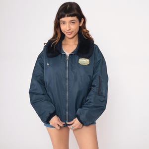 May include: Navy blue bomber jacket with a black faux fur collar. The jacket features a zipper closure, a patch that reads "Oil Well Service Co", and a small embroidered name. The jacket has a ribbed cuff and a pocket on the left sleeve.