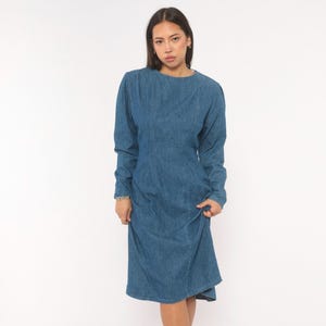 Puede incluir: Un vestido de mezclilla azul de tono medio con mangas largas, cuello redondo y detalles de costuras verticales a lo largo del corpi&ntilde;o. La prenda tiene una cintura ajustada que se transforma en una falda ligeramente acampanada.
