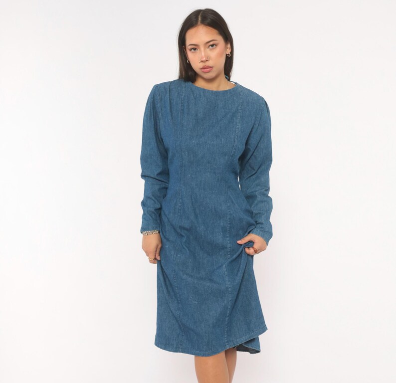 Puede incluir: Un vestido de mezclilla azul de tono medio con mangas largas, cuello redondo y detalles de costuras verticales a lo largo del corpi&ntilde;o. La prenda tiene una cintura ajustada que se transforma en una falda ligeramente acampanada.