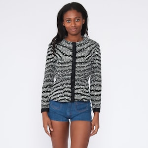 May include: A black and white patterned jacket with a round neckline and long sleeves. The jacket has a button-up front and black trim. The model is wearing blue denim shorts.
