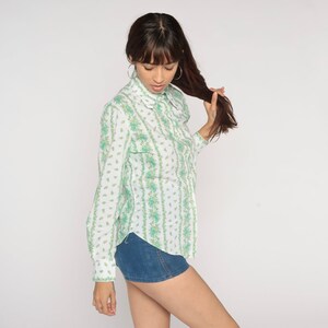 Puede incluir: Una camisa blanca de manga larga con botones y un estampado floral verde y blanco. La camisa tiene cuello y cierre de botones en la parte delantera.