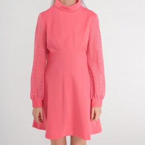 May include: A pink dress with long sleeves. The sleeves are made of a lacy material. The dress has a high neckline and a fitted bodice. The skirt is A-line and falls to just above the knee.