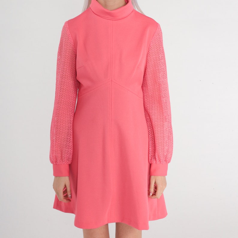 May include: A pink dress with long sleeves. The sleeves are made of a lacy material. The dress has a high neckline and a fitted bodice. The skirt is A-line and falls to just above the knee.