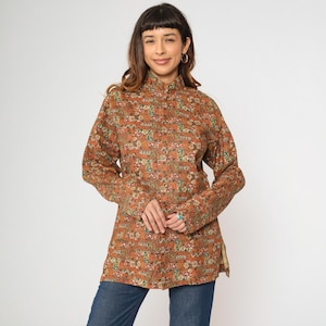 Puede incluir: Una camisa de manga larga con cuello y estampado floral marrón y naranja. La camisa tiene cuello alto y cierre de botones en la parte delantera.