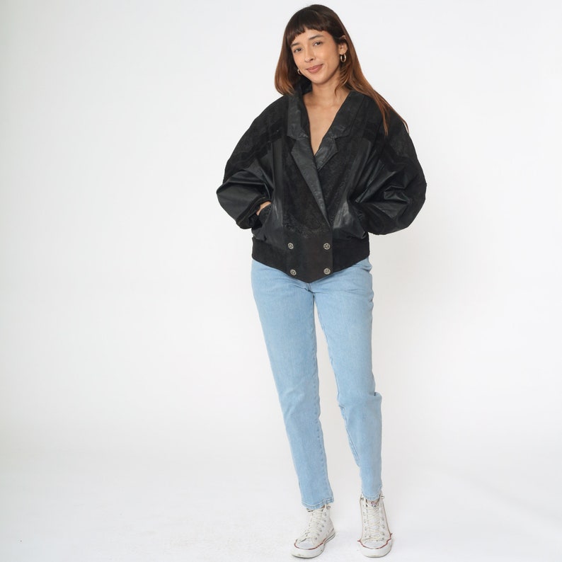 May include: A black leather bomber jacket with a double-breasted closure and a slightly oversized fit. The jacket has a textured surface and is worn over a pair of light blue jeans.
