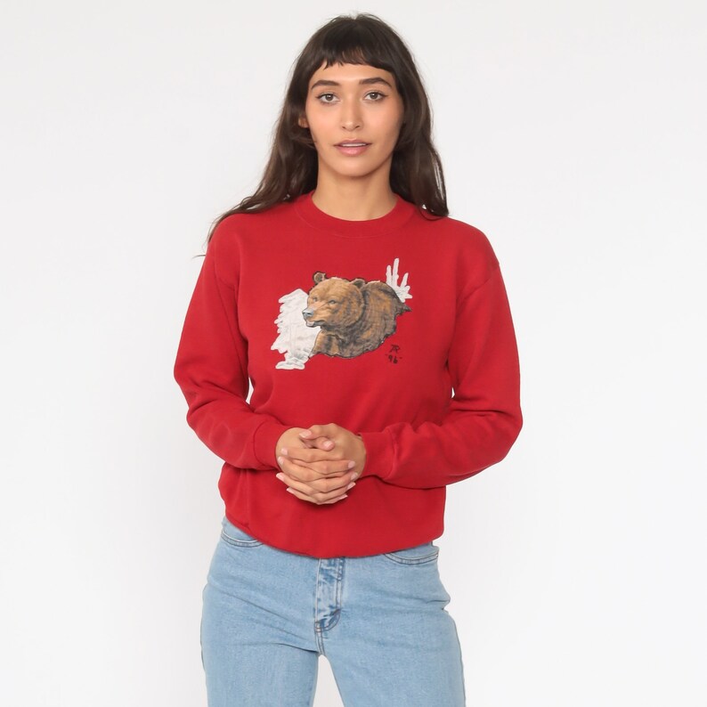 May include: Red sweatshirt with a brown bear graphic on the front. The bear is peeking out from behind a white background.