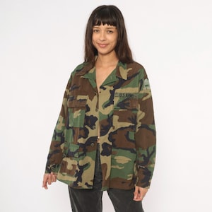 Könnte beinhalten: Jacke mit Camouflage-Muster mit mehreren Taschen und Knopfleiste. Auf der Jacke steht der Text "U.S. ARMY" auf der Brust. Die Jacke ist in Grün-, Braun- und Schwarztönen gehalten.