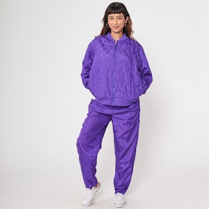 Puede incluir: Un vibrante conjunto de chándal morado. La chaqueta presenta un diseño acolchado con cierre de cremallera y pequeños adornos. Los pantalones a juego tienen un corte holgado. El atuendo se combina con zapatillas blancas, creando una estética deportiva y retro.