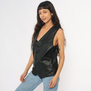 May include: Black leather vest with a V-neck and button closure. The vest has a tailored fit and a small pocket on the front. A classic piece of outerwear.