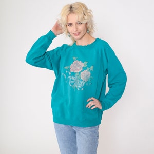 May include: Teal blue sweatshirt with a floral print of pink roses and green leaves. The sweatshirt has a crew neck and long sleeves.