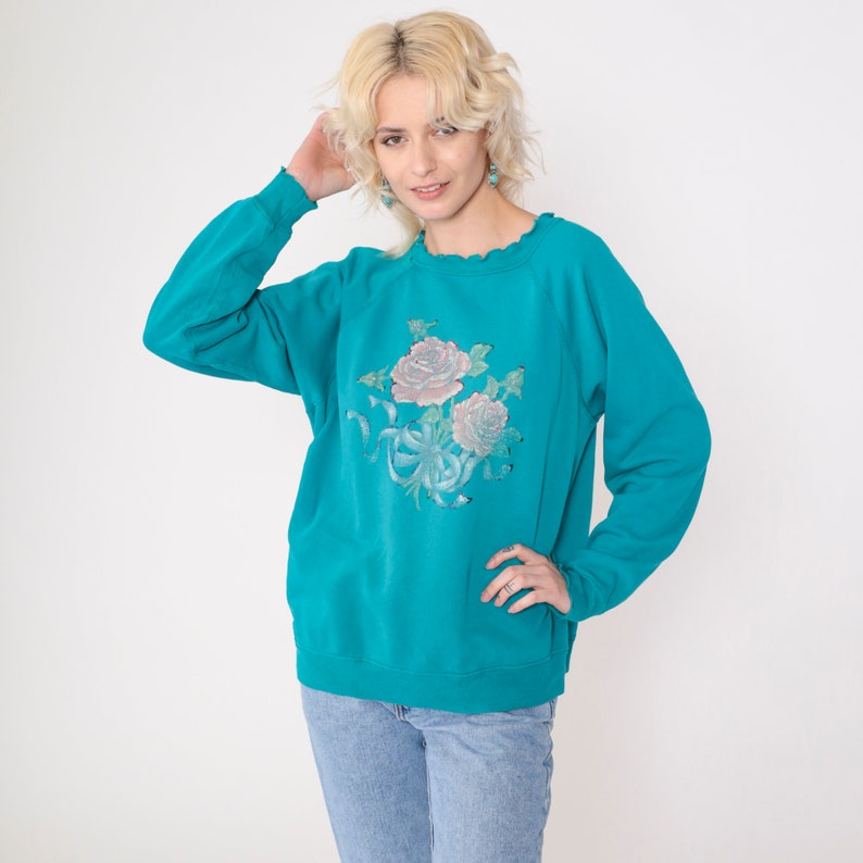 May include: Teal blue sweatshirt with a floral print of pink roses and green leaves. The sweatshirt has a crew neck and long sleeves.