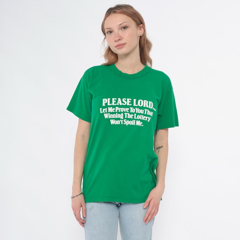 K&ouml;nnte beinhalten: Gr&uuml;nes T-Shirt mit dem Text "PLEASE LORD... Let Me Prove To You That Winning The Lottery Won't Spoil Me." Das Shirt hat einen Rundhalsausschnitt und kurze &Auml;rmel. Die Person tr&auml;gt eine hellblaue Jeans.