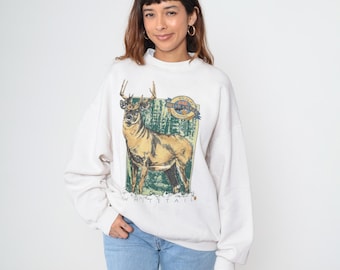 May include: Cream-colored sweatshirt featuring a detailed illustration of a deer with antlers, set against a backdrop of trees. The graphic includes the text "WHITETAIL" and "WILDLIFE NUTRITION". The sweatshirt has a crew neck and long sleeves.