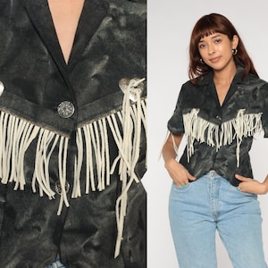 Könnte beinhalten: Dunkelgraue Wildlederjacke mit weißem Fransenbesatz und silbernen herzförmigen Akzenten. Die Jacke hat einen Knopfverschluss und ist im Western-Stil gehalten. Die Fransen verlaufen entlang des Kragens und der Ärmel.