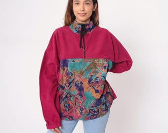 May include: A magenta pullover jacket with a colorful geometric pattern on the front. The jacket has a high collar with a black drawstring and long sleeves. The geometric pattern includes teal, brown, and purple shapes.
