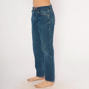 May include: Blue denim jeans with a classic five-pocket design. The jeans have a straight leg cut and a button and zipper closure. The denim has a medium wash with subtle fading and wear marks. The jeans are shown from the side.