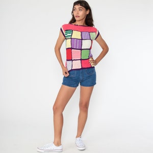 May include: A woman is wearing a pink, multi-colored crochet top with a square pattern. The top has short sleeves and a scoop neck. She is also wearing denim shorts and white sneakers.