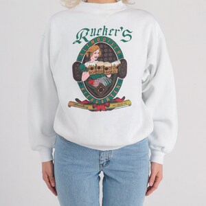 May include: White sweatshirt with a green, red, and yellow graphic print of a woman holding beer steins. The graphic says "Rucker's Altmo&ouml;disch Biergarten Heidelberg est. 1614"