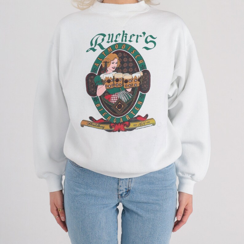 May include: White sweatshirt with a green, red, and yellow graphic print of a woman holding beer steins. The graphic says "Rucker's Altmo&ouml;disch Biergarten Heidelberg est. 1614"