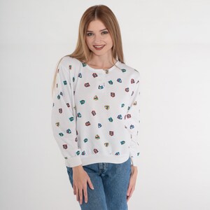May include: White long-sleeve shirt with a button-up neckline and a pattern of colorful shield-like shapes. The shirt is paired with blue jeans. The overall style is casual and comfortable.