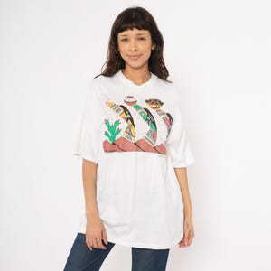 May include: White t-shirt with a colorful graphic print. The design features stylized desert landscape elements, including cacti and abstract shapes in yellow, green, and brown. The shirt has short sleeves and a relaxed fit.