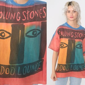 May include: A tie-dye t-shirt with a faded red, orange, blue, and green color scheme. The shirt features a graphic of a face with two eyes and the text "Rolling Stones Voodoo Lounge".