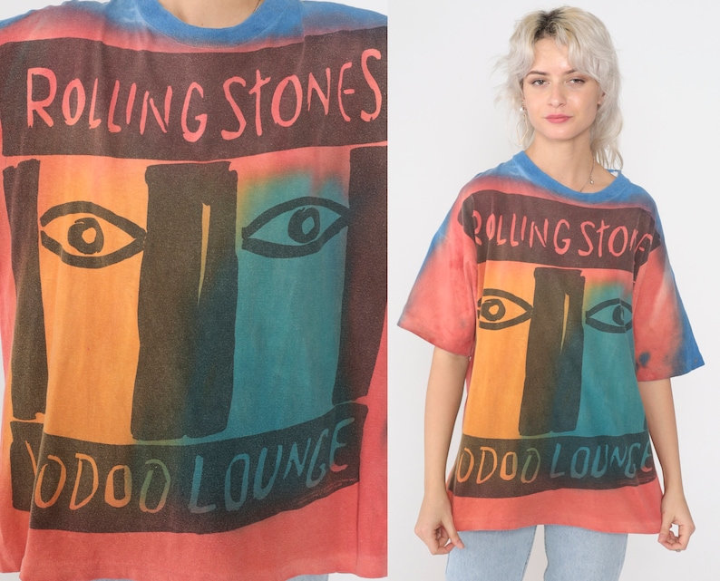 May include: A tie-dye t-shirt with a faded red, orange, blue, and green color scheme. The shirt features a graphic of a face with two eyes and the text "Rolling Stones Voodoo Lounge".